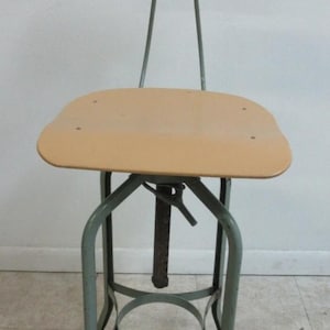 May include: An industrial-style adjustable stool with a light brown seat and backrest. The metal frame is a muted green-gray color. The stool has a circular footrest and a central adjustment mechanism.
