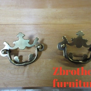 May include: Two ornate brass drawer pulls with a decorative design. The pulls have a curved shape and are attached to a wooden surface.