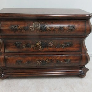 May include: A dark brown wooden dresser with three drawers. The dresser has a curved design and is decorated with floral motifs. The top of the dresser is flat and the base has a decorative trim.