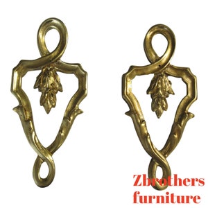 May include: Two gold-toned metal decorative accents with a leaf design and a twisted loop at the top. The accents are in the shape of a heart with a pointed bottom.