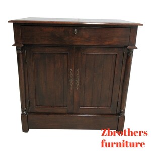May include: A dark brown wooden cabinet with two doors and a single drawer. The cabinet has a simple design with ornate brass handles. The cabinet is sitting on a white background.