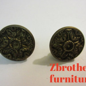 May include: Two ornate gold-toned metal knobs with a floral design. The knobs are round and have a raised center with a smaller circle in the middle. The knobs are likely for furniture.