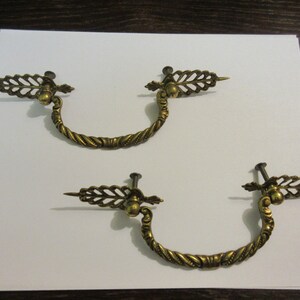 May include: Two antique brass drawer pulls with ornate leaf designs. Each pull has a curved, twisted handle and a decorative knob.