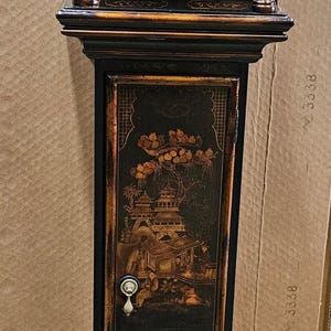 May include: A black grandfather clock with gold accents and a painted scene of a pagoda and flowers. The clock face has Roman numerals and a gold background.