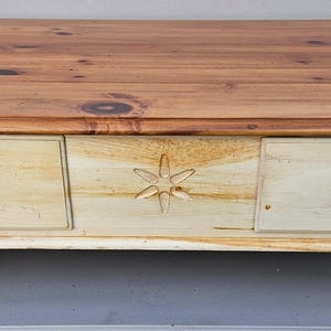 May include: A rectangular coffee table with a light brown wooden top and a cream-colored base. The base features three drawers with round knobs and decorative star carving. The table rests on four rounded feet.
