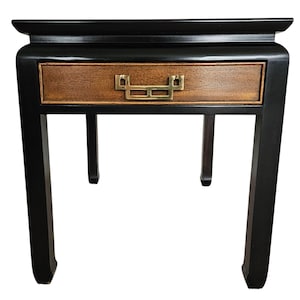 May include: Black wooden side table with a single drawer. The drawer is made of light brown wood and has a brass handle. The table has four black legs and a black top.