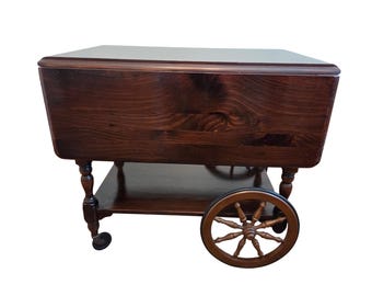 May include: A dark brown wooden tea cart with a rectangular top and a lower shelf. The cart has decorative turned legs, a spoked wheel, and small caster wheels. The wood grain is visible.