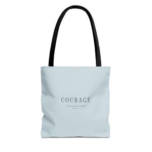 May include: A light blue tote bag with black handles. The front features the word COURAGE in a dark serif font, followed by the text "the Lord thy God is with thee" and "JOSHUA 1:9" in a smaller, matching dark font centered on the bag.