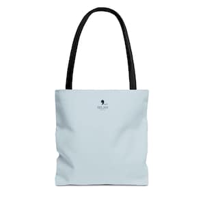May include: A light blue tote bag with two black shoulder straps. The front of the bag features a small dark blue logo consisting of a stylized abstract shape above the text SELAH in a clean, sans-serif font, centered near the top.