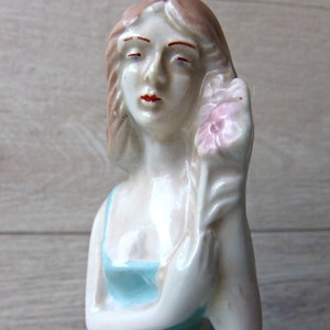 Puede incluir: Figura de porcelana vintage de una mujer sosteniendo una flor rosa. La figura est&aacute; pintada con un vestido azul claro y un tono de piel p&aacute;lido. La mujer tiene labios rojos y cabello casta&ntilde;o.