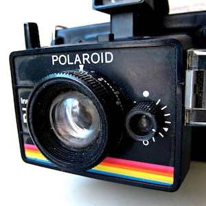 May include: A black Polaroid instant camera with the brand name in white lettering. The camera has a lens, viewfinder, and control dials. A colorful rainbow stripe accents the bottom edge. The camera is a vintage model.