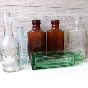 Puede incluir: Colección de botellas de vidrio antiguas de varias formas y colores. Incluye botellas transparentes, ámbar y verdes con texto en relieve. Algunas botellas tienen etiquetas, otras son lisas. Un tema de botellas de farmacia o medicina vintage.