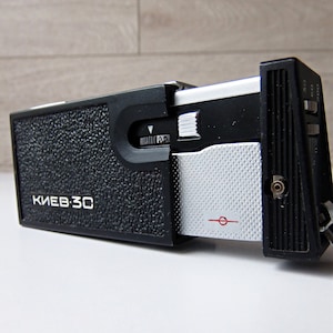 May include: A black and silver Kiev 30 camera. The camera has a textured black body with the text "KIEV 30" in white. It features a silver and black lens and a dial with settings. The camera is a vintage item.
