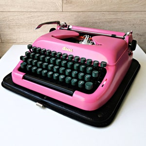 以下が含まれることがあります： 黒いキーボードと緑色のキーを備えた、ヴィンテージのピンク色のタイプライター。 タイプライターは黒いケースの上にあります。 「Olivetti」の文字がタイプライターに表示されています。