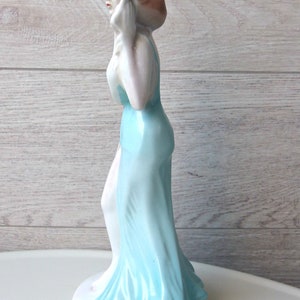 Puede incluir: Figura de porcelana de una mujer con un vestido azul claro y fluido. La figura tiene una tez p&aacute;lida, con labios rojos y cabello casta&ntilde;o. La figura est&aacute; sobre una base blanca.