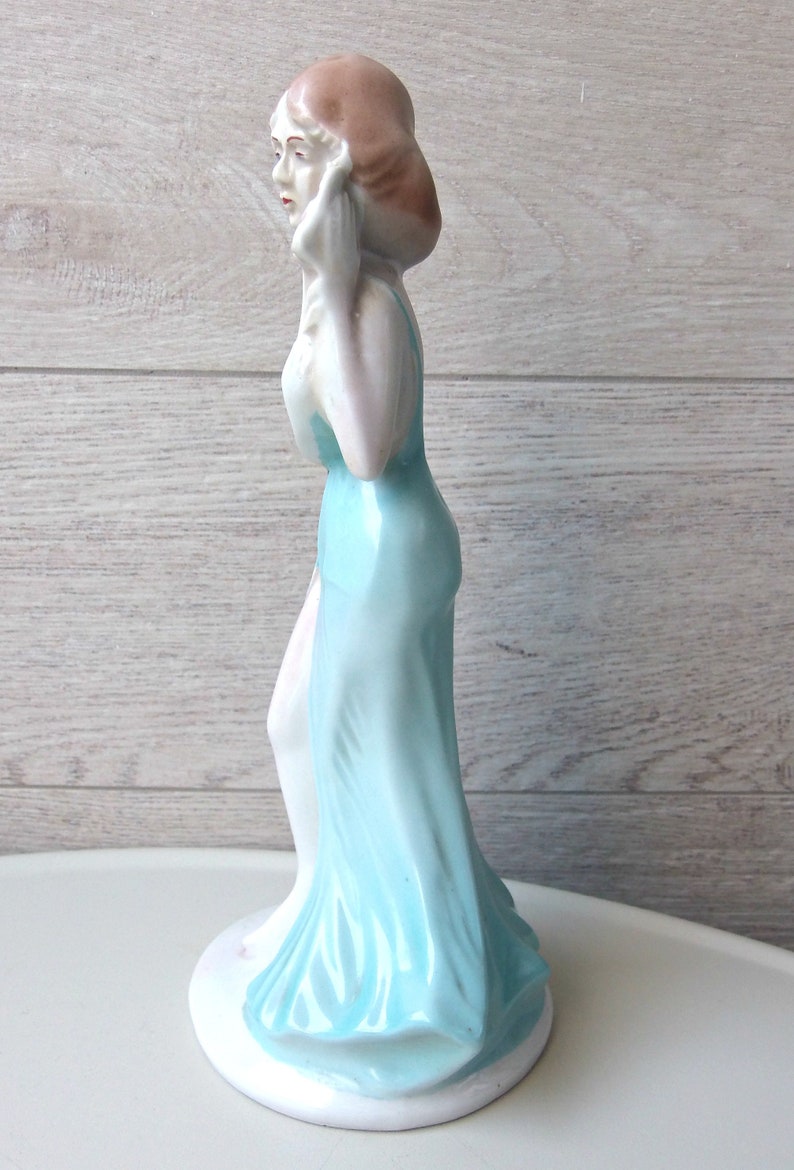 Puede incluir: Figura de porcelana de una mujer con un vestido azul claro y fluido. La figura tiene una tez p&aacute;lida, con labios rojos y cabello casta&ntilde;o. La figura est&aacute; sobre una base blanca.