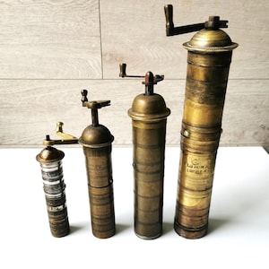 Può includere: Sono presentati quattro antichi macinapepe in ottone di diverse dimensioni. I macinapepe hanno una forma cilindrica con una manovella. L'ottone ha una calda patina invecchiata. I macinapepe sono disposti in fila, mostrando le loro diverse altezze e design.
