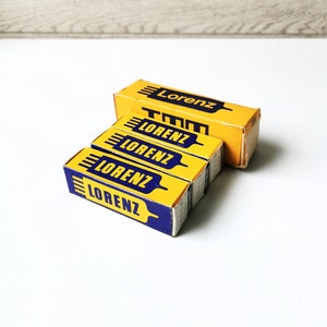 Puede incluir: Varias cajas amarillas y azules de Lorenz están apiladas sobre una superficie blanca. Las cajas tienen la palabra "Lorenz" impresa en una fuente azul oscuro y en negrita. Las cajas son rectangulares y parecen ser vintage o retro.