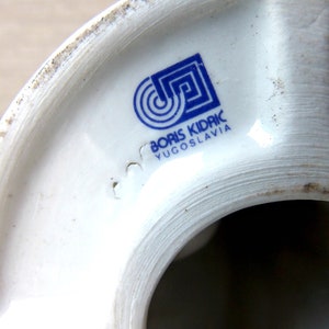 Puede incluir: Primer plano de un objeto de cer&aacute;mica blanca con un logotipo y texto azules. El logotipo presenta un dise&ntilde;o geom&eacute;trico con c&iacute;rculos conc&eacute;ntricos y un cuadrado. El texto dice "BORIS KIDRIC YUGOSLAVIA".