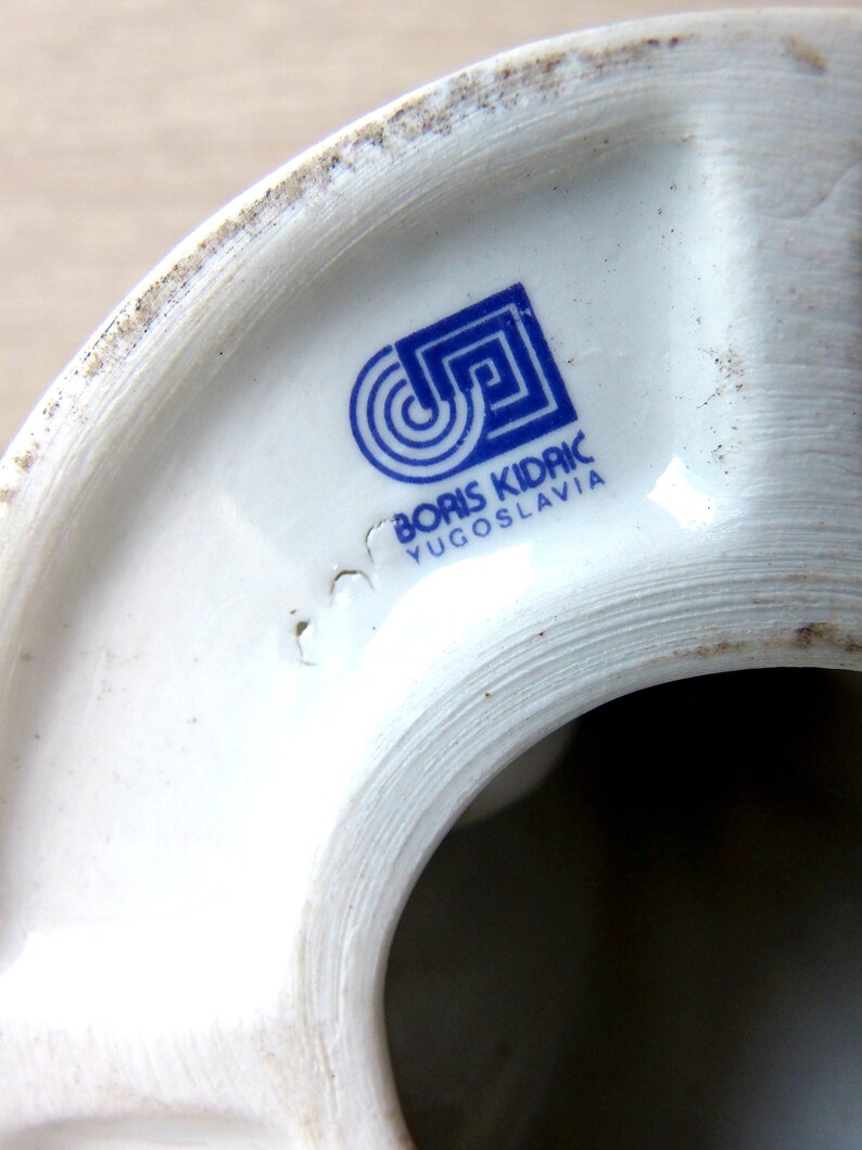 Puede incluir: Primer plano de un objeto de cer&aacute;mica blanca con un logotipo y texto azules. El logotipo presenta un dise&ntilde;o geom&eacute;trico con c&iacute;rculos conc&eacute;ntricos y un cuadrado. El texto dice "BORIS KIDRIC YUGOSLAVIA".