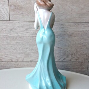 Puede incluir: Figura de cer&aacute;mica de una mujer con un vestido azul claro y fluido. La figura se ve desde atr&aacute;s, mostrando el elegante dise&ntilde;o del vestido y el cabello casta&ntilde;o de la mujer. La figura est&aacute; sobre una base blanca.