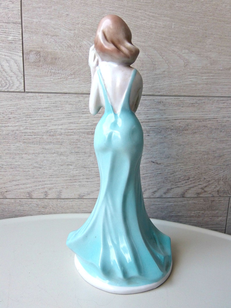 Puede incluir: Figura de cer&aacute;mica de una mujer con un vestido azul claro y fluido. La figura se ve desde atr&aacute;s, mostrando el elegante dise&ntilde;o del vestido y el cabello casta&ntilde;o de la mujer. La figura est&aacute; sobre una base blanca.