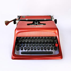 May include: A vintage red typewriter with black keys and a red return key. The typewriter has a sleek design with the text "nimu Studio 44" visible. The keys are arranged in a QWERTY layout.