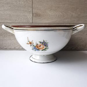 May include: White enamelware bowl with two handles and a pedestal base. The bowl features a floral design with orange, blue, and purple flowers. The rim is brown with gold trim. A vintage kitchenware item.