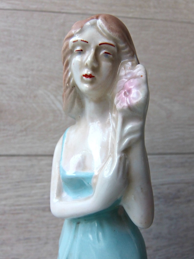Puede incluir: Figura de porcelana vintage de una mujer sosteniendo una flor rosa. La figura est&aacute; pintada con un vestido azul claro y un tono de piel p&aacute;lido. La mujer tiene labios rojos y cabello casta&ntilde;o.