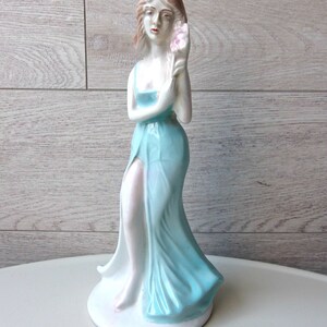 Puede incluir: Figura de cer&aacute;mica de una mujer con un vestido azul claro y fluido, sosteniendo una flor rosa. La figura tiene una base blanca y est&aacute; sobre un fondo claro de vetas de madera. La mujer tiene el pelo casta&ntilde;o claro y est&aacute; en una pose elegante.