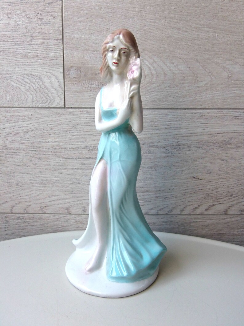 Puede incluir: Figura de cer&aacute;mica de una mujer con un vestido azul claro y fluido, sosteniendo una flor rosa. La figura tiene una base blanca y est&aacute; sobre un fondo claro de vetas de madera. La mujer tiene el pelo casta&ntilde;o claro y est&aacute; en una pose elegante.