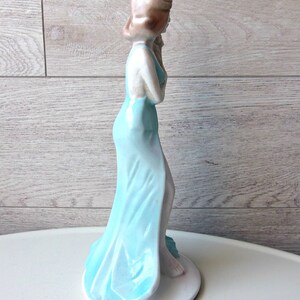 Puede incluir: Figura de cer&aacute;mica de una mujer con un vestido azul claro y fluido. La figura tiene el pelo casta&ntilde;o claro y est&aacute; de pie sobre una base circular blanca. El vestido tiene un efecto degradado, que pasa de un tono azul m&aacute;s oscuro a uno m&aacute;s claro.