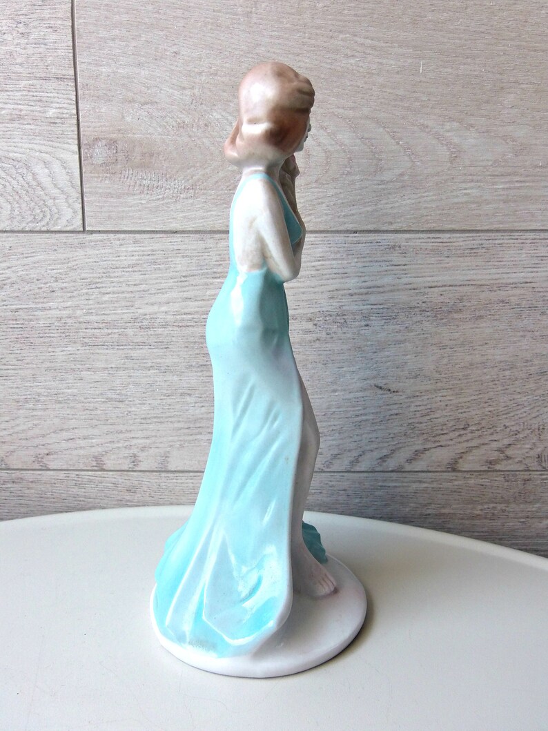 Puede incluir: Figura de cer&aacute;mica de una mujer con un vestido azul claro y fluido. La figura tiene el pelo casta&ntilde;o claro y est&aacute; de pie sobre una base circular blanca. El vestido tiene un efecto degradado, que pasa de un tono azul m&aacute;s oscuro a uno m&aacute;s claro.