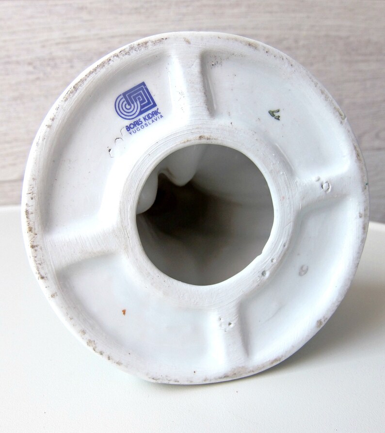Puede incluir: Vista inferior de una figura de cer&aacute;mica blanca con base circular y abertura central. La base presenta un logotipo azul con el texto "BORIS KIDRIC YUGOSLAVIA". La figura tiene un acabado liso y brillante.