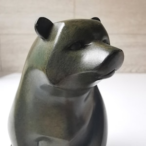 May include: A bronze-coloured bear figurine with a smooth, glossy finish. The bear is depicted in a seated position, with its head turned slightly to the right. The sculpture has small, rounded ears and a detailed snout, showcasing a blend of naturalistic and stylized elements.