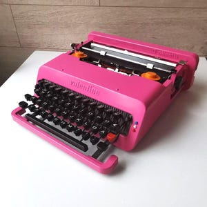 May include: A bright pink Olivetti Valentine typewriter with black keys and orange accents. The word "valentine" is printed on the front. The typewriter sits on a white surface.