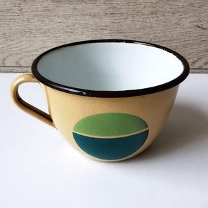 May include: A vintage enamelware cup with a tan exterior, a black rim, and a white interior. The cup features a green and teal oval design on the side and a curved handle. The cup is likely used for drinking beverages.