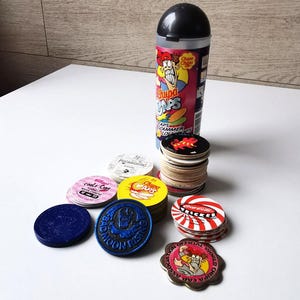 May include: A selection of Chupa Caps, comprising a stack of circular game pieces and a container. The caps display diverse designs and text, with a blue, yellow, and red colour palette. The container is cylindrical with a black top and vibrant graphics.