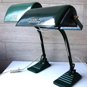 May include: Two vintage green desk lamps with the word "HORAX" on the shade. The lamps have a curved black arm and a rectangular base. The lamps are on a white surface.