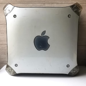 Op de afbeelding: Een zilveren Apple-computer met een donkerblauw Apple-logo. De hoeken zijn versterkt met doorzichtig plastic. De computer heeft een vierkante vorm en is waarschijnlijk een vintage model.