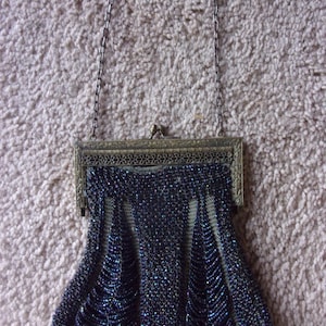 May include: A small, dark blue beaded handbag featuring a metallic, antique-style frame with intricate filigree detailing. The bag is suspended by a thin, matching metal chain handle and displays a textured, scalloped pattern of tiny beads.