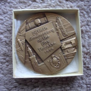 May include: A circular bronze-toned commemorative medallion housed in a square box. The medallion features relief carvings of architectural elements and a central plaque inscribed with Fiftieth Anniversary of the Fox Theatre 1979.