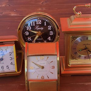 May include: A collection of vintage clocks in various shapes and sizes. The clocks feature gold-toned frames and include a round clock with a black face, a square clock with a white face, and a rectangular clock with an orange face.
