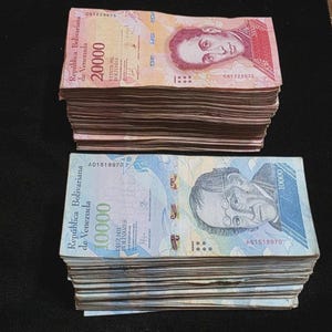 May include: Two thick stacks of Venezuelan currency. The top stack consists of pink 20000 Bolivares notes featuring a portrait. The bottom stack contains blue 10000 Bolivares notes with a portrait of a man wearing glasses. Both stacks are rectangular.
