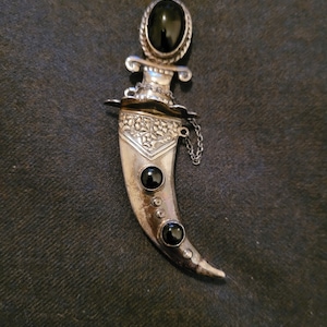 Peut inclure: Une broche en forme de poignard en argent avec des pierres d'onyx noir. Le poignard a un design détaillé et une chaîne attachée au manche.