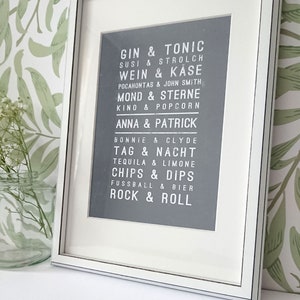 Puede incluir: Una impresi&oacute;n enmarcada con fondo gris y texto blanco. El texto est&aacute; en una fuente de estilo vintage y enumera pares de palabras, como "Gin & Tonic", "Susi & Strolch", "Wein & Kase", "Mond & Sterne", "Anna & Patrick", "Tag & Nacht", "Chips & Dips", "Rock & Roll".