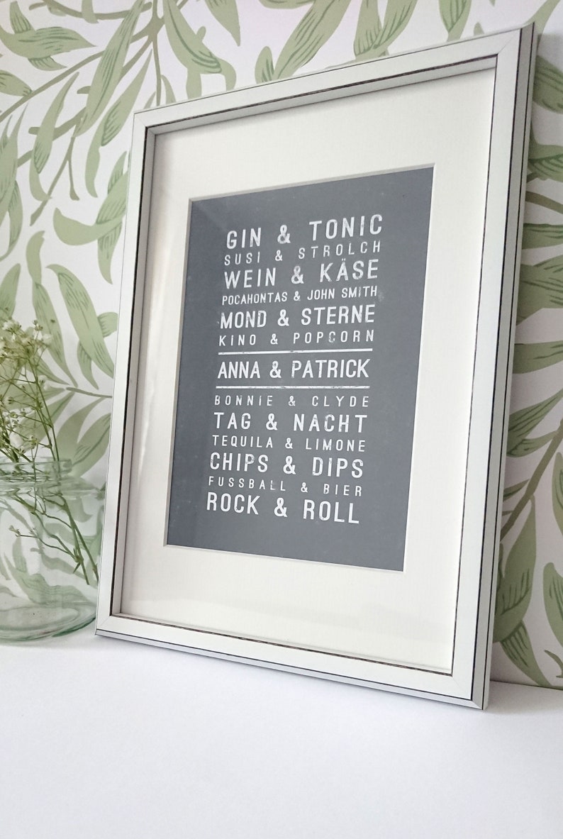 Puede incluir: Una impresi&oacute;n enmarcada con fondo gris y texto blanco. El texto est&aacute; en una fuente de estilo vintage y enumera pares de palabras, como "Gin & Tonic", "Susi & Strolch", "Wein & Kase", "Mond & Sterne", "Anna & Patrick", "Tag & Nacht", "Chips & Dips", "Rock & Roll".