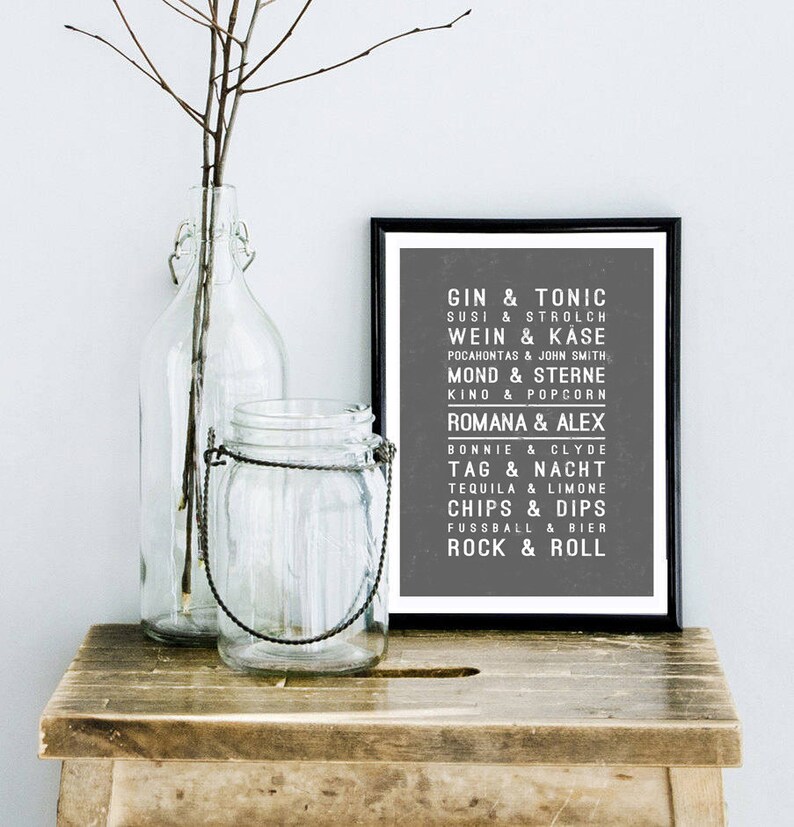 May include: A black and white framed print with a gray background and white text. The text lists various pairs of words, such as "Gin & Tonic", "Susi & Strolch", "Wein & K&auml;se", "Pocahontas & John Smith", "Mond & Sterne", "Kino & Popcorn", "Romana & Alex", "Bonnie & Clyde", "Tag & Nacht", "Tequila & Limone", "Chips & Dips", "Fussball & Bier", and "Rock & Roll".