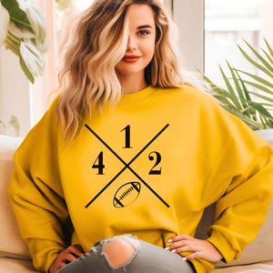 May include: A yellow sweatshirt with a black graphic design. The design features the numbers 1, 2, and 4, with a football in the center. The sweatshirt is a solid yellow color.