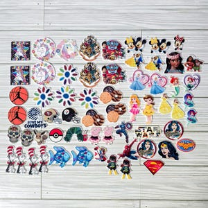 May include: A collection of colorful, decorative charms featuring various designs. Includes unicorns, cartoon characters, sports logos, and floral patterns. The charms are arranged on a light wood-grain surface.